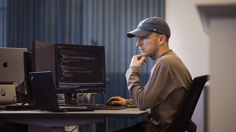 En mand i kasket arbejder koncentreret ved en computer med flere skærme, der viser programmeringskode.